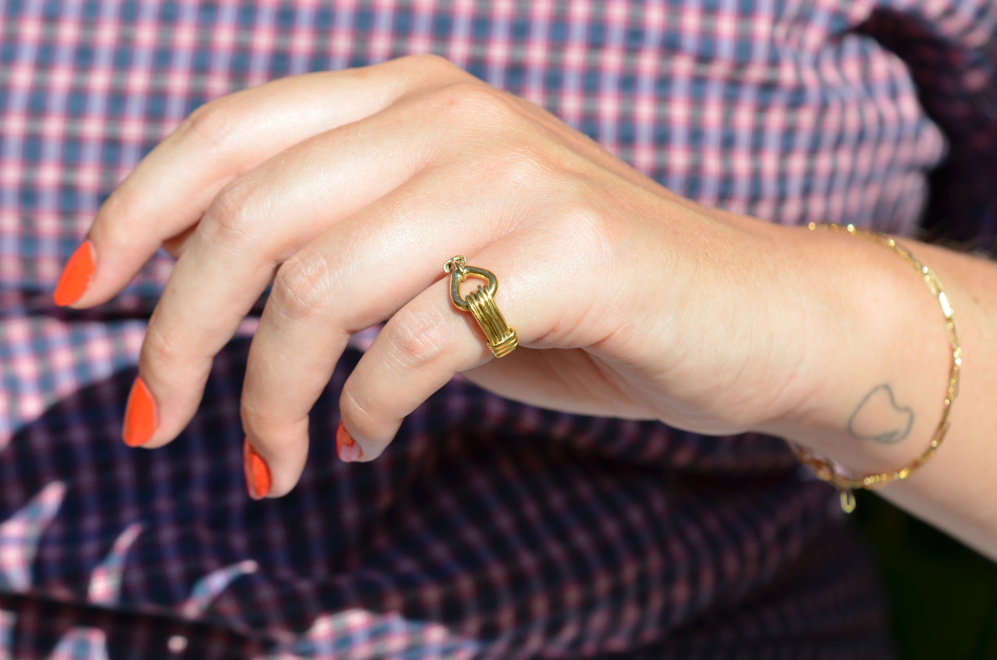 A medium-close view of the ring on a Caucasian model's left pinky finger. Her nail polish is an orangey-red, and in the background her blue and red gingham blouse is visible.