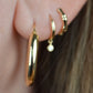 the featured hoops are shown on a caucasian model's first lobe piercing. slightly out of focus are a snug gold hoop with dangling diamond in the second piercing, a snug gold hoop stylized as bamboo in the third piercing.