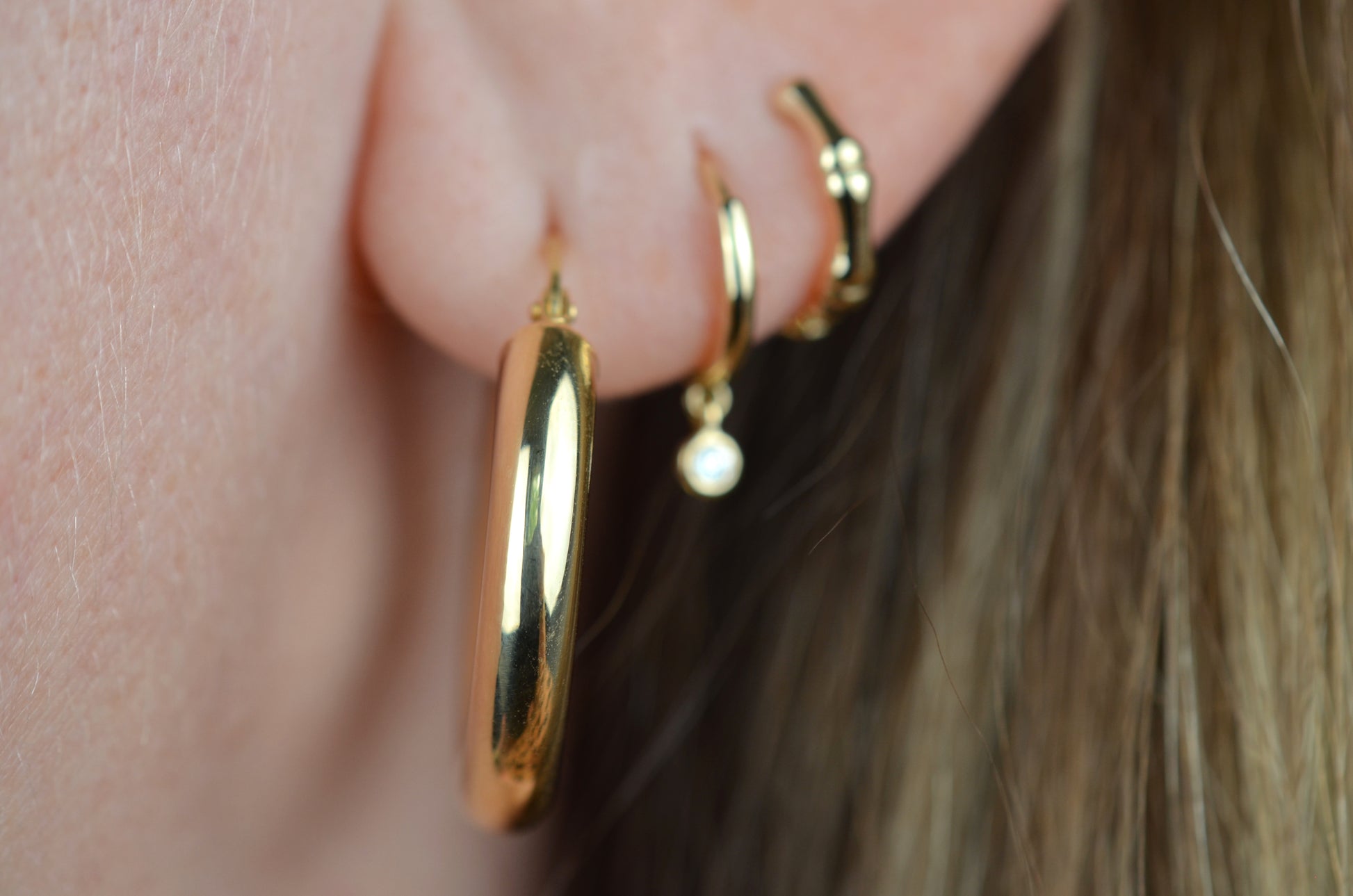 the featured hoops are shown on a caucasian model's first lobe piercing. slightly out of focus are a snug gold hoop with dangling diamond in the second piercing, a snug gold hoop stylized as bamboo in the third piercing.