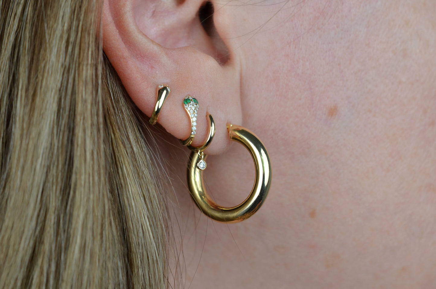 on a caucasian model, the featured hoops are shown in a first lobe piercing and fit loosely. also shown in piercings going up the lobe are a snug gold hoop with a dangling diamond, a snug gold hoop stylized as a snake with pavé diamonds and emerald eyes, and a snug gold hoop stylized as a snake with diamond eyes.