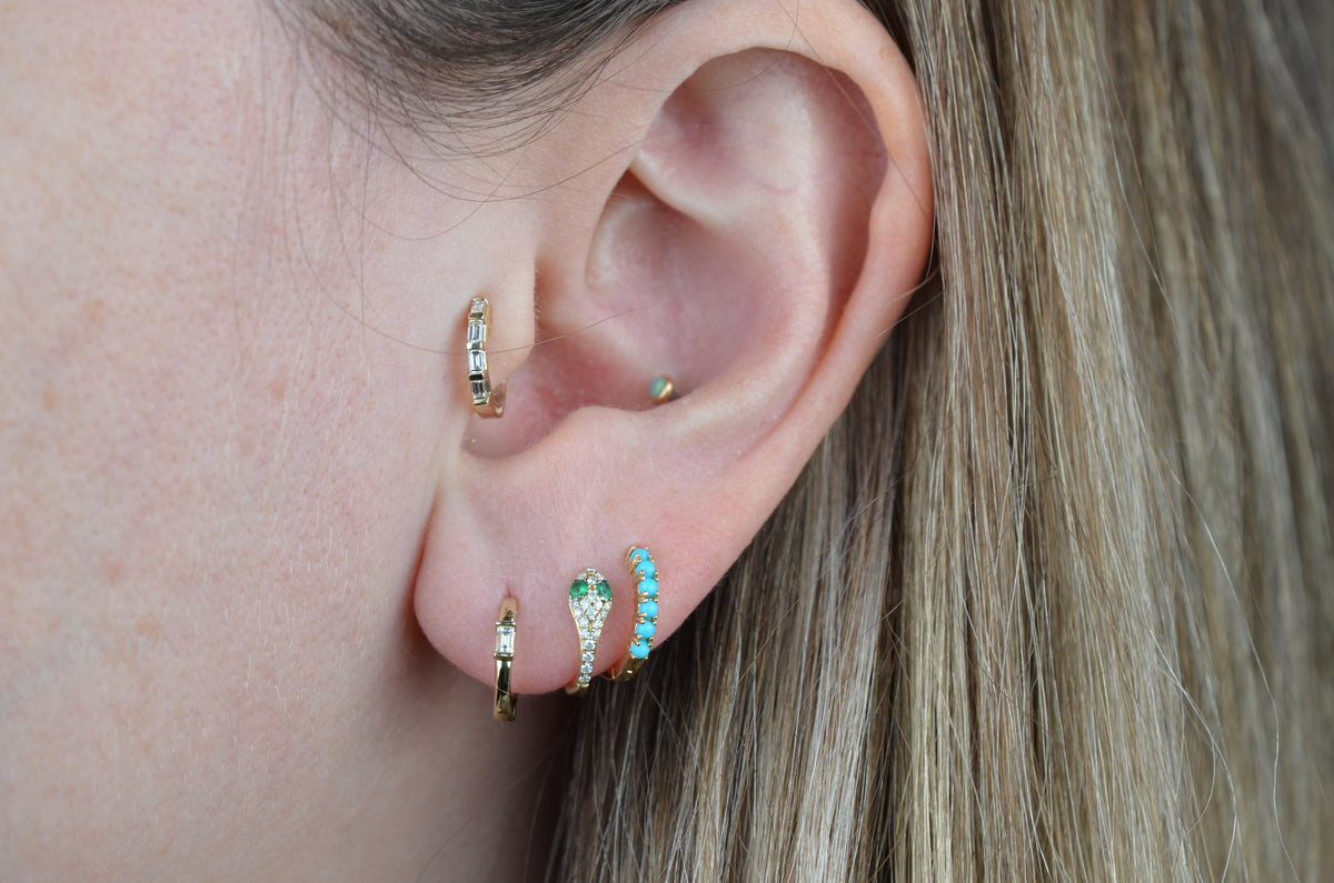a caucasian model wears the featured earring in a third lobe piercing, where the fit is snug. additionally visible are a gold and baguette diamond small hoop in the first piercing, a pavé diamond and emerald-eyed snake small hoop in the second piercing, a multi-stone baguette diamond hoop in the tragus piercing, and a small opal stud in the conch piercing.