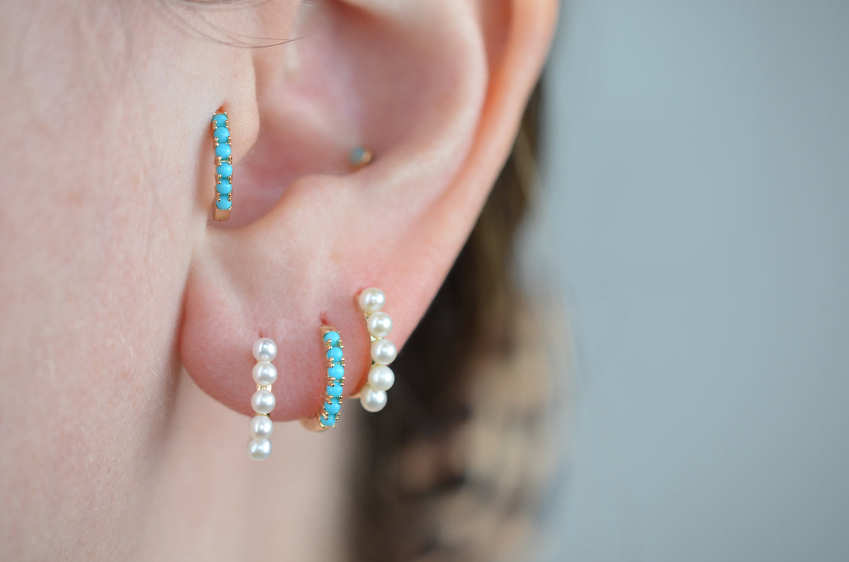 a caucasian model wears the featured turquoise small hoop in both the second lobe piercing and tragus piercing. in the first and third lobe piercing are a pair of gold and pearl small hoop earrings.