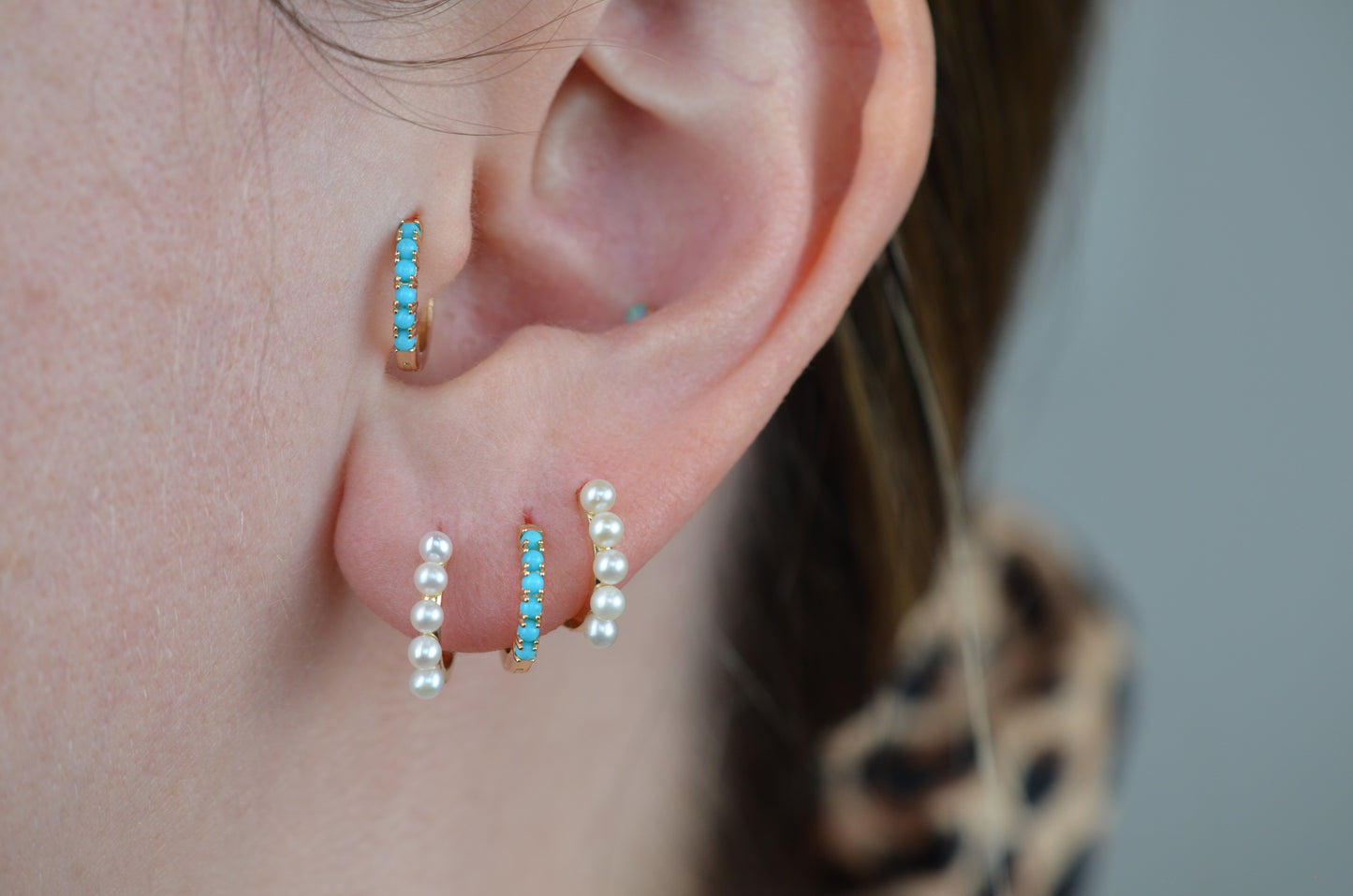 a caucasian model wears the featured turquoise small hoop in both the second lobe piercing and tragus piercing. in the first and third lobe piercing are a pair of gold and pearl small hoop earrings.