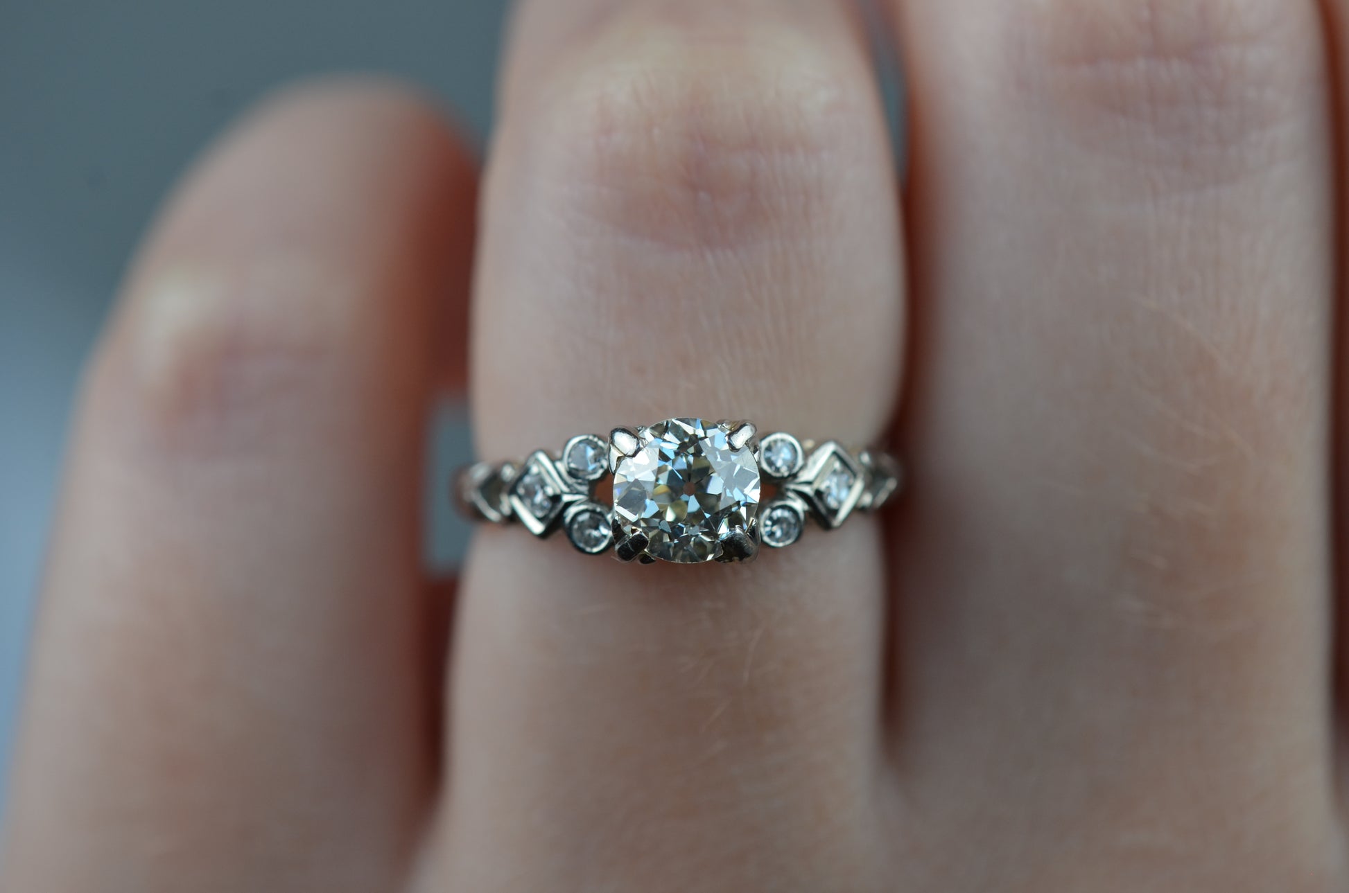 a head-on macro of the ring on a caucasian model's left ring finger. the ring is in crisp focus while the hand is slightly blurry. lighting is cool indoor lighting, making the ring and primary diamond appear whiter.