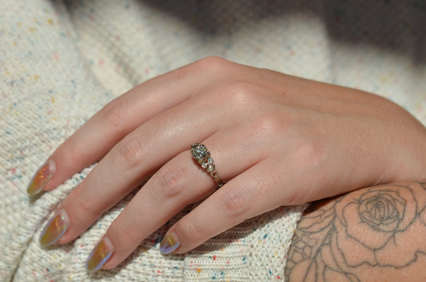 the ring is styled on a caucasian model's left ring finger in direct sunlight. the shoulder details are visible, as well as the model's shimmering gold fingernails and a floral arm tattoo.