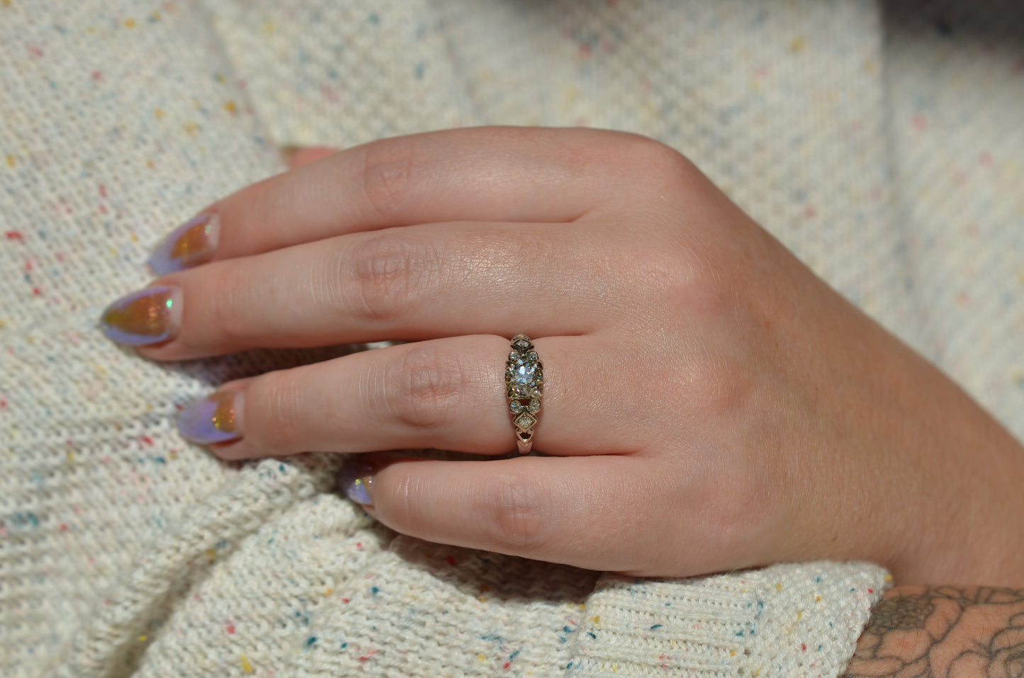 the ring is styled on a caucasian model's left ring finger in direct sunlight. the shoulder details are visible, as well as the model's shimmering gold fingernails and a floral arm tattoo.